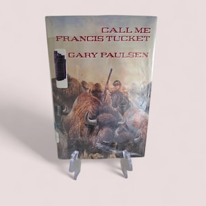 May include: A hardcover book titled "CALL ME FRANCIS TUCKET" by Gary Paulsen. The cover features a painting of bison and a person on horseback. The book is displayed on a clear plastic stand.