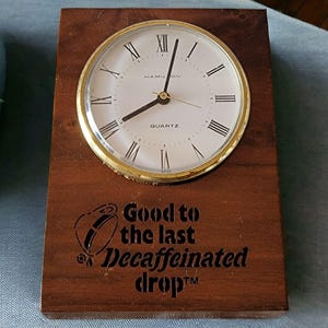 Puede incluir: Un reloj de madera con una esfera dorada y agujas negras. El reloj está montado en una placa de madera con el texto "Good to the last Decaffeinated drop™" grabado en ella.