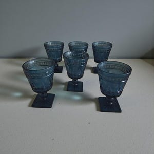 May include: Six blue-tinted vintage water goblets. Each glass features a square base, a stem, and a detailed bowl with a decorative pattern. The glasses are arranged on a white surface, with a gray background.