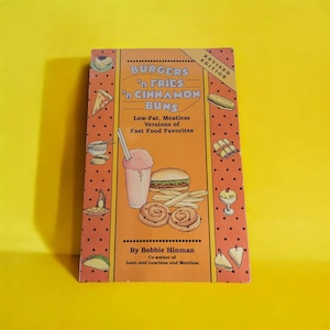 May include: A cookbook titled "Burgers 'n Fries 'n Cinnamon Buns" with illustrations of fast food items. The cover is orange with a dotted border and features a burger, fries, a milkshake, and cinnamon buns. The book is a revised edition.
