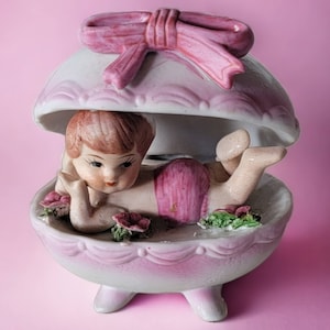 May include: A porcelain figurine of a girl laying inside a pink and white egg-shaped shell with a pink bow on top.