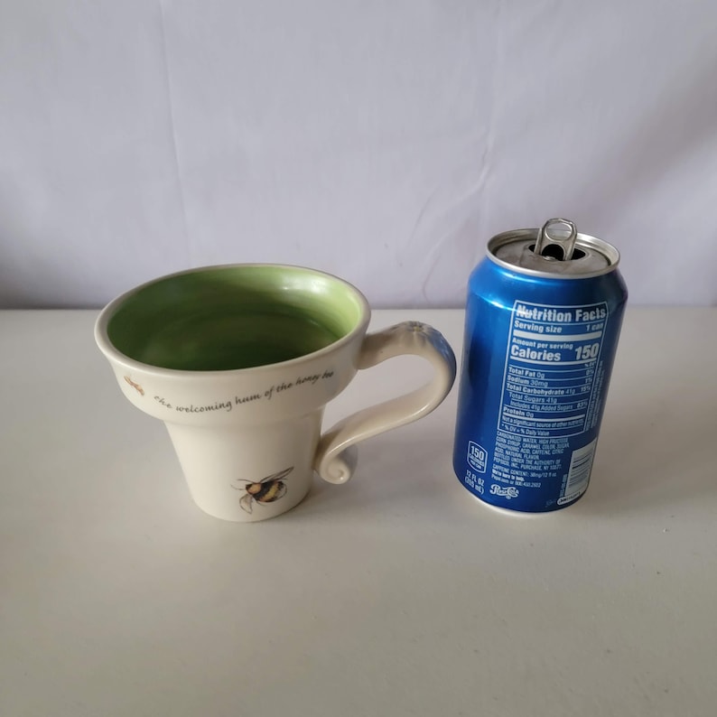 May include: Cream-colored ceramic mug with a green interior and a bee illustration. The mug has a curved handle and the text "the welcoming hum of the honey bee". A blue soda can is next to the mug.