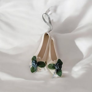 May include: A pair of white porcelain ballet shoes with green leaves and blue flowers. The shoes are tied together with a white ribbon and are hanging from a white ribbon.