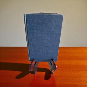 Puede incluir: Un libro azul oscuro se muestra en un pequeño soporte decorativo. El libro tiene una cubierta texturizada y está ligeramente inclinado. El soporte es de color oscuro y tiene un diseño único y estilizado. Los artículos están sobre una superficie de madera.