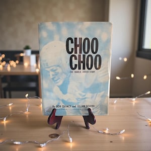 Puede incluir: Un libro antiguo titulado "CHOO CHOO The Charlie Justice Story" se exhibe sobre una mesa de madera. La portada del libro presenta una imagen en blanco y negro de un jugador de fútbol americano y el título en letras negras en negrita. El libro está sostenido por un pequeño soporte de madera rojo oscuro.