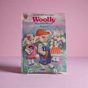 Puede incluir: Un libro infantil titulado "Woolly Has Two Dozen Sisters!" presenta ilustraciones de ovejas de dibujos animados con monos, vestidos y sombreros. La portada del libro tiene un fondo rosa y morado con el texto "Itsy-Bitsies Storybooks".