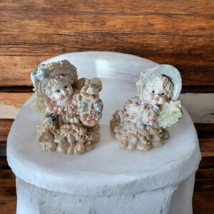 May include: Two small porcelain figurines of children wearing hats and dresses. The figurines are sitting on a white surface and are decorated with flowers and other details.