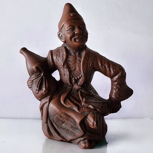 May include: A brown ceramic figurine of a person wearing a hat and holding a jug. The figurine is sitting down with one leg bent and the other leg straight out.