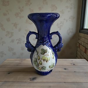 May include: A cobalt blue ceramic vase with a flared top and two curved handles. The vase features a central oval design with a botanical illustration of green and yellow berries and leaves. The vase is placed on a wooden surface.