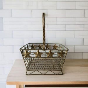 May include: A decorative metal basket with a rectangular shape and a handle. The basket is made of wire mesh and features gold-colored star accents around the top. The handle is a twisted chain. The basket is sitting on a light-colored wooden surface.