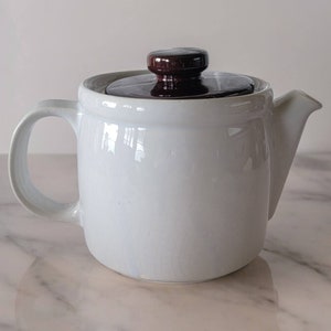 May include: White ceramic teapot with a brown lid. The teapot has a rounded body and a spout on the side. The teapot is sitting on a white marble surface.