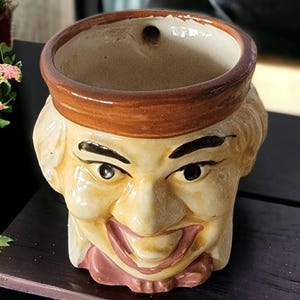 May include: A ceramic planter shaped like a smiling man's head with a brown hat. The planter has a hole in the top for drainage.