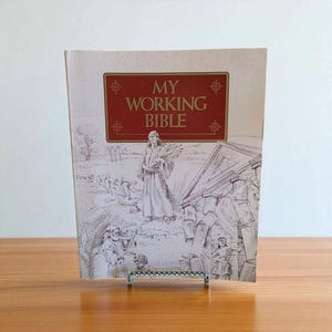 Peut inclure: Un livre intitulé "MY WORKING BIBLE" avec une bannière rectangulaire rouge sur la couverture. La couverture présente une illustration d'une personne portant des gerbes de blé, avec d'autres personnages en arrière-plan. Le livre est présenté sur un support en métal argenté.