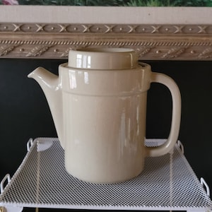 Puede incluir: Una tetera de cerámica beige con tapa y asa. La tetera está sobre una rejilla de alambre blanca.