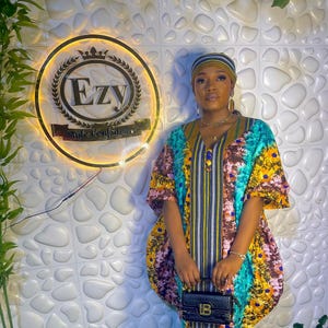 May include: A colorful, patterned dress with a matching head wrap. The dress features a mix of teal, yellow, and brown hues. A small black purse is held in front. The background includes a sign with the text "Ezy Style Confidence."