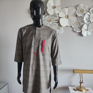 May include: A brown plaid two-piece suit on a mannequin. The shirt features a pocket with a red zigzag detail. The suit has a classic collar and half sleeves. The trousers are straight-legged.