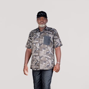 Può includere: Un uomo indossa una camicia a maniche corte con stampa mimetica e un berretto nero con la scritta "BLACK" in lettere bianche. Indossa anche pantaloncini di jeans blu scuro e scarpe nere.