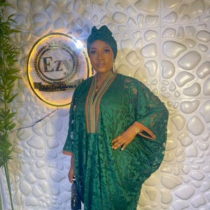 May include: A dark green, flowing dress with a matching head wrap. The dress features a floral pattern and orange trim along the neckline and sleeves. A small black handbag is held in the hand.