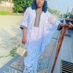 May include: A white lace dress with floral patterns, a matching head wrap, and a gold handbag. The dress has a V-neck with brown and gold trim. The person is wearing gold-colored shoes.