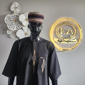 May include: Black traditional African attire, including a short-sleeved tunic with embroidered details and a matching cap. The tunic features gold-colored embroidery on the chest and pocket. The cap has a striped pattern in shades of brown and blue.