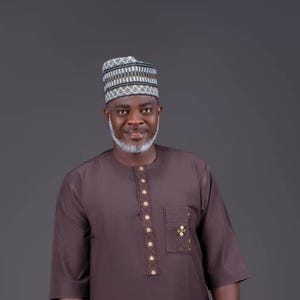 May include: A man wearing a brown traditional outfit, including a long-sleeved shirt and matching pants. He is also wearing a patterned cap. The outfit has gold-colored buttons and a pocket with a decorative emblem.