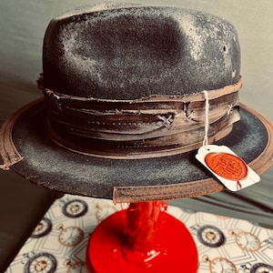 Può includere: Un cappello fedora grigio antracite scuro con una finitura usurata e invecchiata. Il cappello presenta un'ampia fascia in pelle marrone a strati con cuciture incrociate visibili e un'etichetta bianca attaccata da un cordino, con un sigillo in cera rosso.