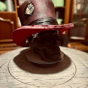 Puede incluir: Un sombrero de copa rojo oscuro con acabado desgastado, que presenta una banda negra y un parche con costuras visibles en el lateral. El sombrero está colocado sobre un modelo de cráneo negro, con el borde inferior de color rojo brillante.