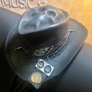 May include: A black fedora hat featuring a distressed finish, a black and white paisley patterned band, silver-toned metal spikes, and a dark chain. Attached to the hat are two white square tags and a gold-coloured wax seal with a logo.