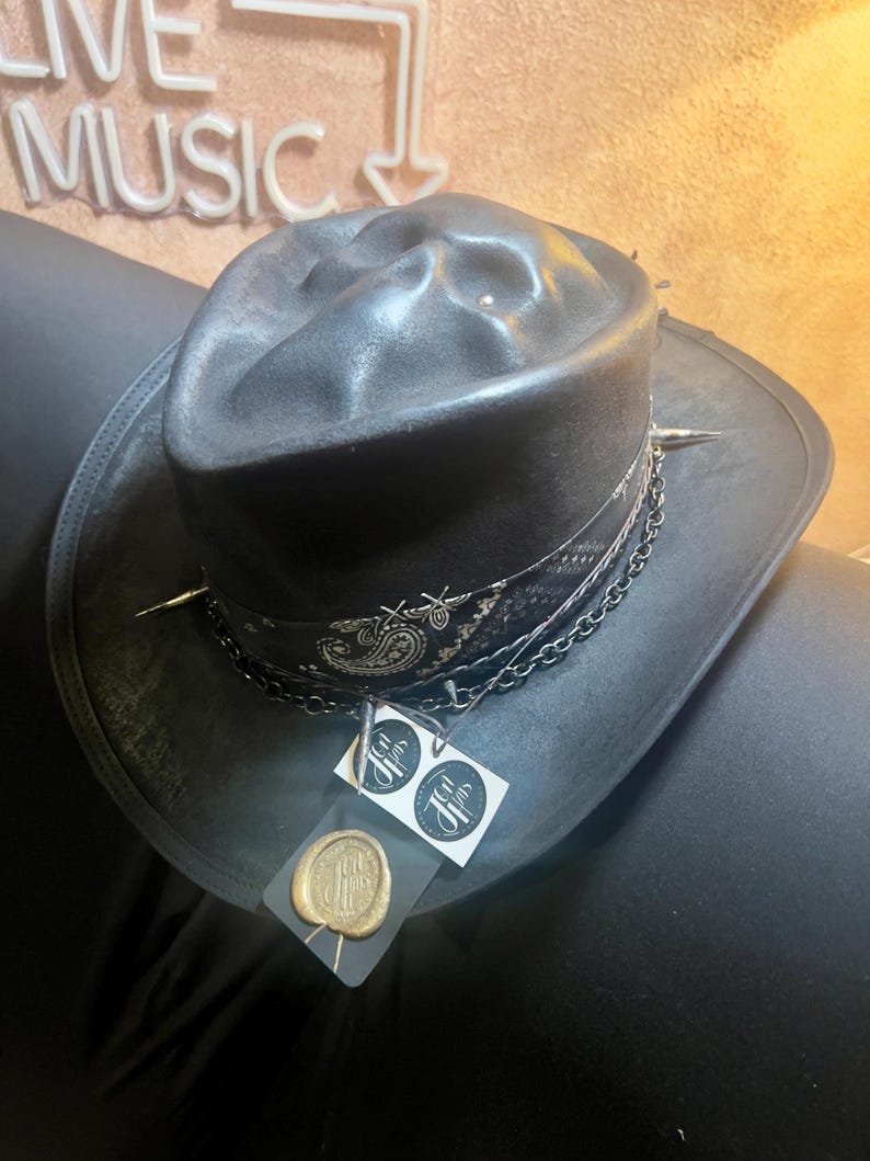 May include: A black fedora hat featuring a distressed finish, a black and white paisley patterned band, silver-toned metal spikes, and a dark chain. Attached to the hat are two white square tags and a gold-coloured wax seal with a logo.