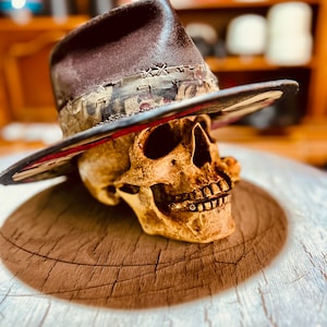 May include: A dark brown fedora hat with a distressed, multi-coloured patterned band featuring visible cross-stitch detailing. The hat is positioned atop a realistic human skull model, showcasing a wide brim and a structured, pinched crown design.