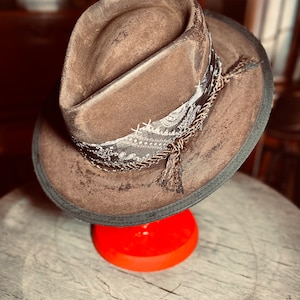 Könnte beinhalten: Ein brauner Fedora-Hut mit abgenutztem Finish, der ein schwarz-weißes Band mit Paisley-Muster um die Krone gewickelt hat, akzentuiert durch dünne Metallketten und eine kleine braune Quaste, die an der Seite der Hutkrempe herabhängt.