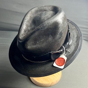 May include: A distressed dark grey fedora hat with a black ribbon band, a thin silver chain, and a small silver sword charm. A white rectangular tag with a red wax seal featuring the text "Jen Has" is attached to the side of the hat band.