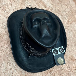 May include: A black hat featuring a moulded skull design on the crown, a black bandana-style band with white patterns, and silver-toned spikes. A black tag with the text "Ten Jhas" and a circular gold-coloured wax seal emblem are attached to the side.