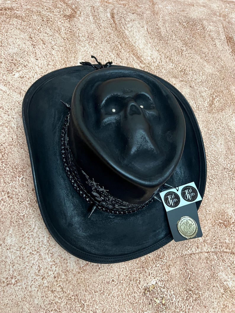 May include: A black hat featuring a moulded skull design on the crown, a black bandana-style band with white patterns, and silver-toned spikes. A black tag with the text "Ten Jhas" and a circular gold-coloured wax seal emblem are attached to the side.