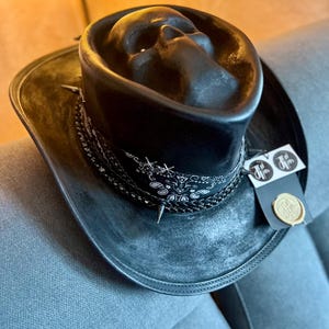 May include: A black hat featuring a moulded skull shape on the crown, a black and white paisley bandana band, and a braided trim with small metal spikes. A rectangular tag with a circular logo and a gold wax seal is attached to the side of the brim.