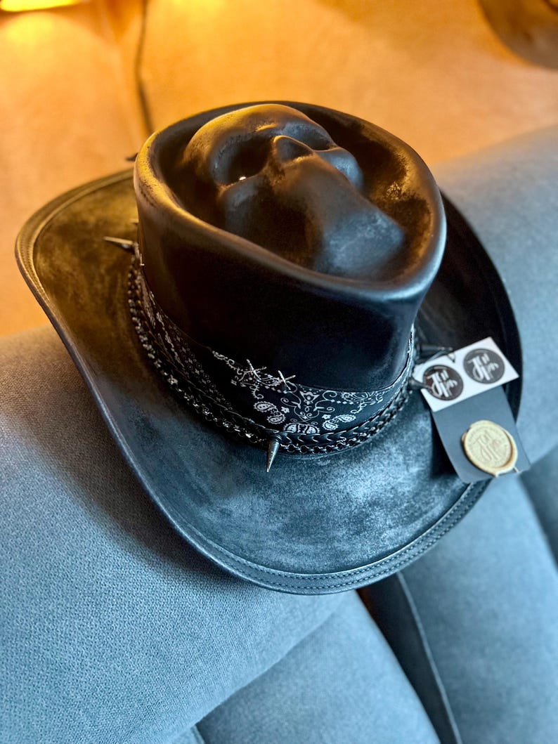 May include: A black hat featuring a moulded skull shape on the crown, a black and white paisley bandana band, and a braided trim with small metal spikes. A rectangular tag with a circular logo and a gold wax seal is attached to the side of the brim.
