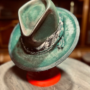 May include: A distressed green fedora hat with a dark, weathered finish. The crown features a deep center crease and a pinched front. A decorative band wraps around the base, featuring a black and white patterned fabric with braided trim.