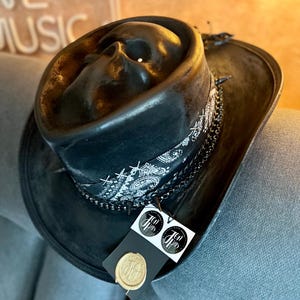 May include: A black hat featuring a moulded skull on the crown, wrapped with a black and white paisley bandana, a dark metal chain, and small spikes. A black tag with two circular white logos and a gold wax seal is attached to the hat brim.