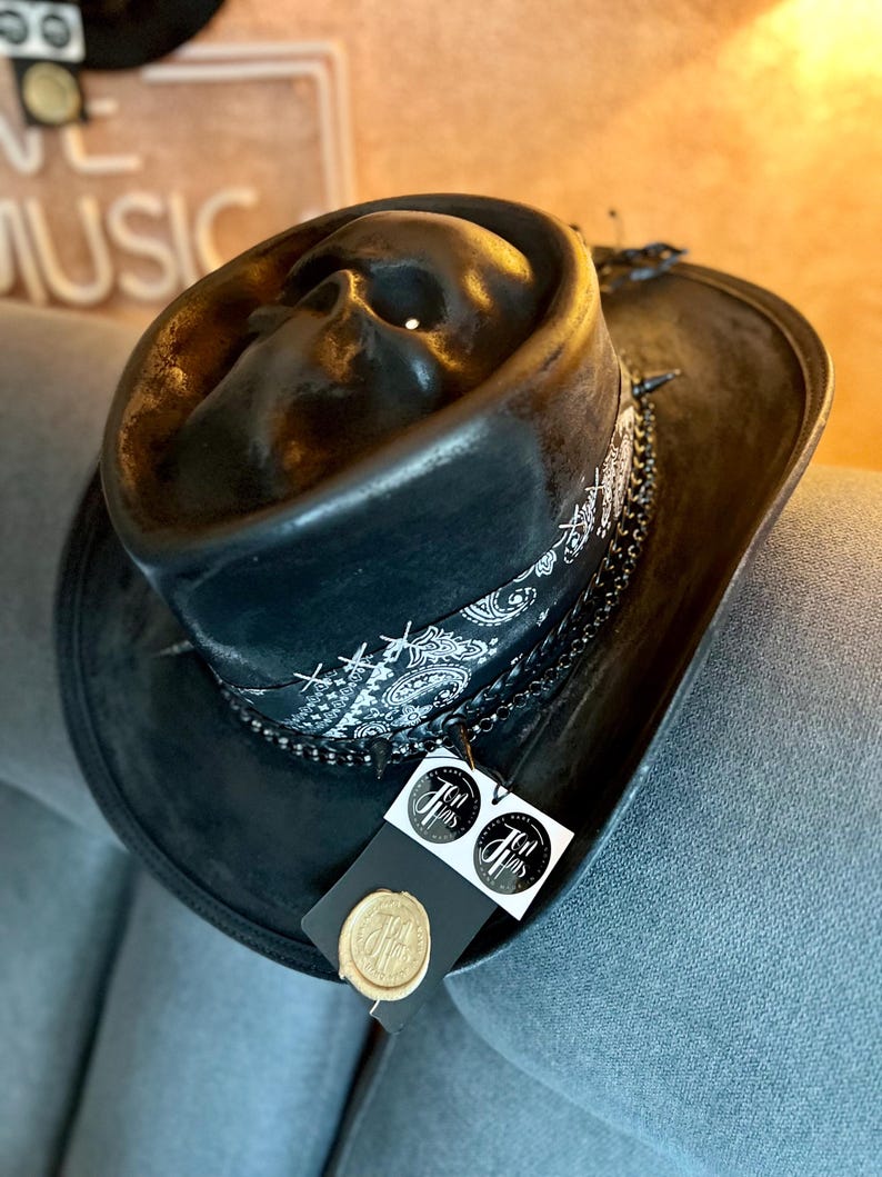 May include: A black hat featuring a moulded skull on the crown, wrapped with a black and white paisley bandana, a dark metal chain, and small spikes. A black tag with two circular white logos and a gold wax seal is attached to the hat brim.