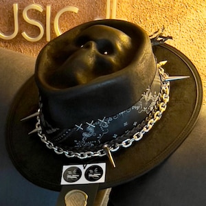 Puede incluir: Un sombrero fedora negro con una corona en forma de calavera, una banda de pañuelo negro con patrones de cachemira blancos, púas de metal plateado y una cadena plateada. Una etiqueta rectangular con dos logotipos y un sello está unida al ala.