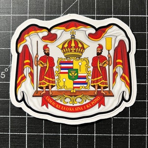 May include: A white sticker with a red, yellow, and blue coat of arms. The coat of arms features two figures holding staffs, a crown, and a shield with a Hawaiian flag. The text "Ua Mau Ke Ea O Ka Aina I Ka Pono" is written on a red ribbon.