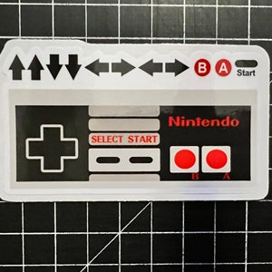 May include: A white sticker with a black and white illustration of a Nintendo Entertainment System controller. The controller has a black background with white buttons and a white Nintendo logo. The sticker has a white border.