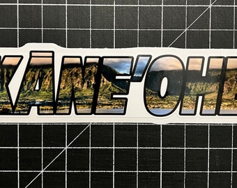 May include: A white sticker with black outlining featuring the text "KÄNE'ÖHE" in bold, black letters. The letters are filled with a scenic image of a mountain range and a body of water.
