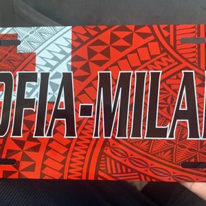 May include: A red, white, and blue license plate with a Polynesian design and the name "SOFIA-MILANI" printed in black.