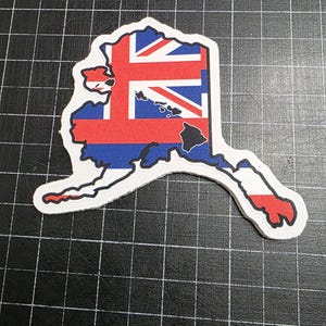 May include: A sticker in the shape of the state of Alaska, featuring a design that incorporates the Union Jack and the state flag colors. The sticker has a white border and is set against a black grid background.
