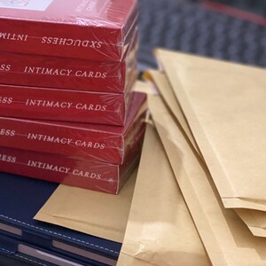 K&ouml;nnte beinhalten: Ein Stapel roter Boxen mit dem Text "INTIMACY CARDS" auf jeder Box. Die Boxen sind in Klarsichtfolie verpackt und liegen auf einem Stapel brauner Umschl&auml;ge.