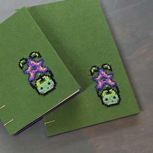 K&ouml;nnte beinhalten: Zwei gr&uuml;ne Notizb&uuml;cher mit einem Kreuzstichmuster eines Cartoon-Charakters. Die Figur hat einen lila seesternartigen Kopf und einen gr&uuml;nen K&ouml;rper. Die Notizb&uuml;cher haben lila Kanten und liegen auf einer dunklen Holzoberfl&auml;che.