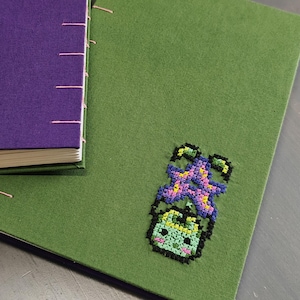 K&ouml;nnte beinhalten: Zwei handgefertigte Journale. Eines ist lila mit rosa N&auml;hten, das andere ist gr&uuml;n mit einem Kreuzstichmuster einer l&auml;chelnden Pflanze. Die Journale liegen auf einer grauen Oberfl&auml;che.