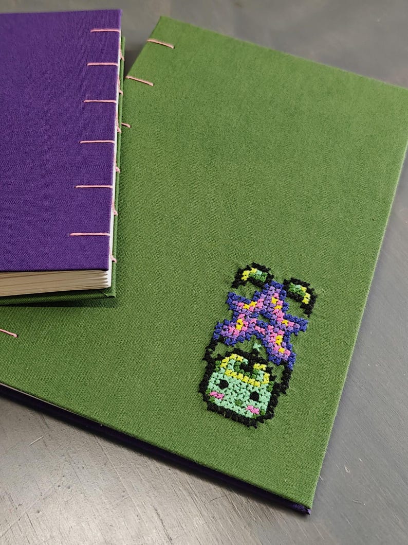 K&ouml;nnte beinhalten: Zwei handgefertigte Journale. Eines ist lila mit rosa N&auml;hten, das andere ist gr&uuml;n mit einem Kreuzstichmuster einer l&auml;chelnden Pflanze. Die Journale liegen auf einer grauen Oberfl&auml;che.