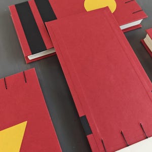 K&ouml;nnte beinhalten: Mehrere rechteckige Notizb&uuml;cher mit roten Einb&auml;nden, die schwarze Streifen und gelbe geometrische Formen wie einen Kreis und ein Dreieck aufweisen. Die Notizb&uuml;cher sind mit sichtbaren schwarzen F&auml;den an den R&auml;ndern gebunden und enthalten wei&szlig;e Papierseiten.